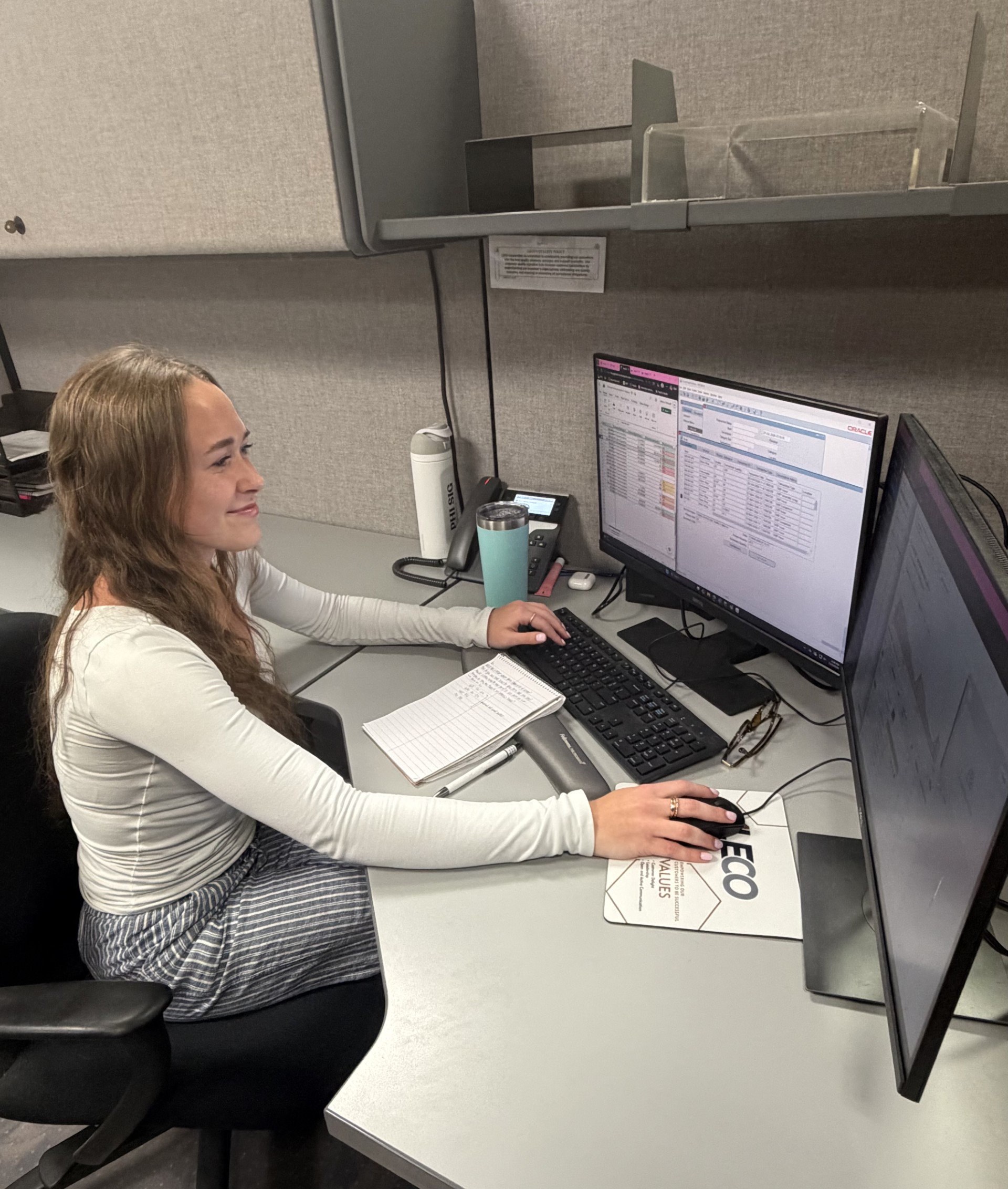 A young woman sits at a desk with two monitors and smiles at what is on her screen.