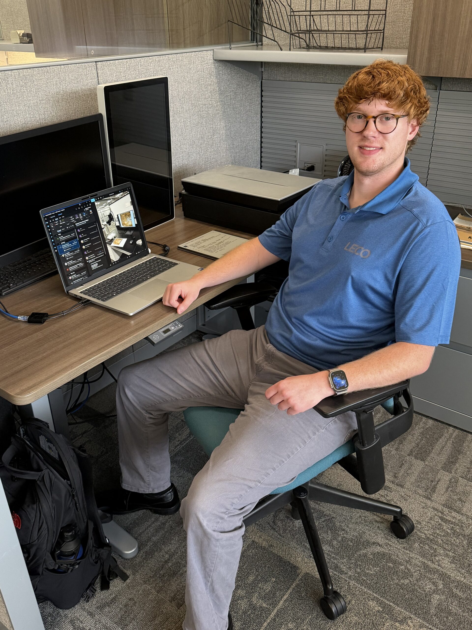 A smiling red-haired young man at a desk with a laptop and 2 monitors.