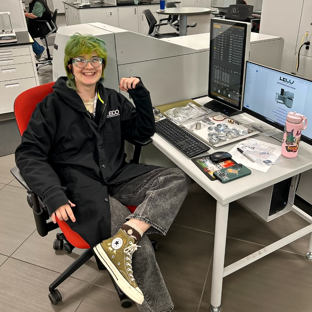 A young, green-haired woman with glasses smiles at the camera from a small table with a few monitors and notebooks.